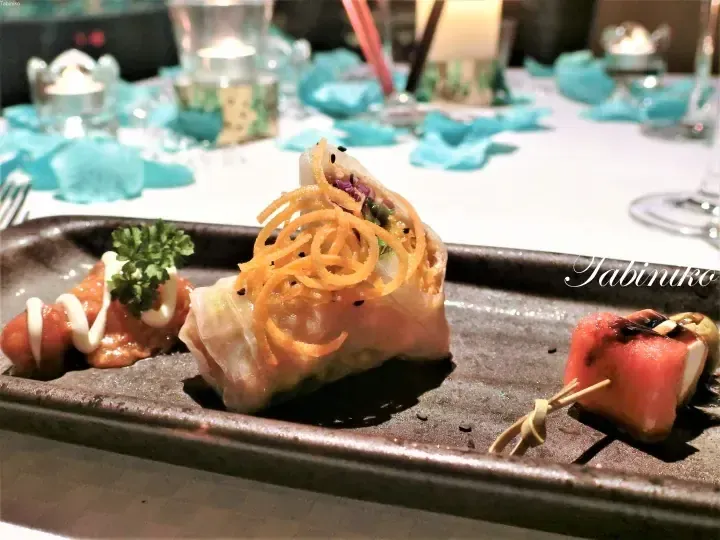 A close-up of an elegant appetizer platter featuring a spring roll garnished with crispy noodles, served on a dark rectangular plate.
