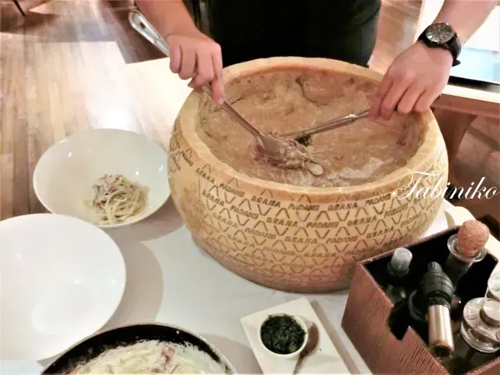 Chef serving pasta from a hollowed Grana Padano cheese wheel at a dining table.