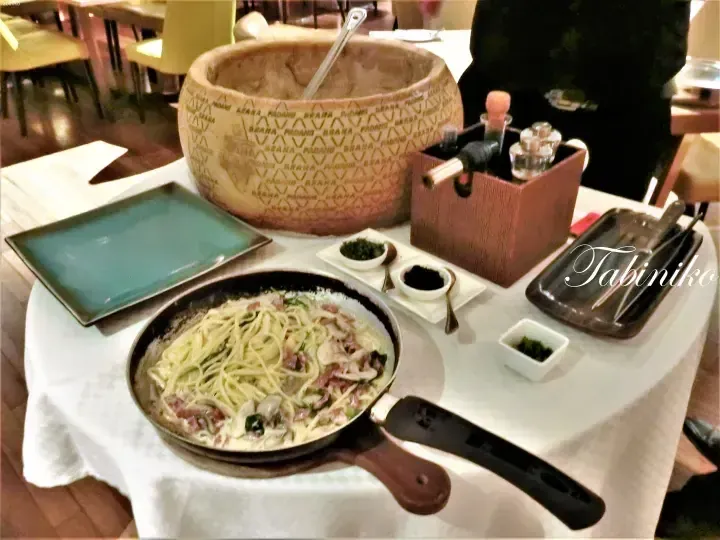 Creamy pasta served in a skillet alongside a large Grana Padano cheese wheel and condiments at a restaurant table