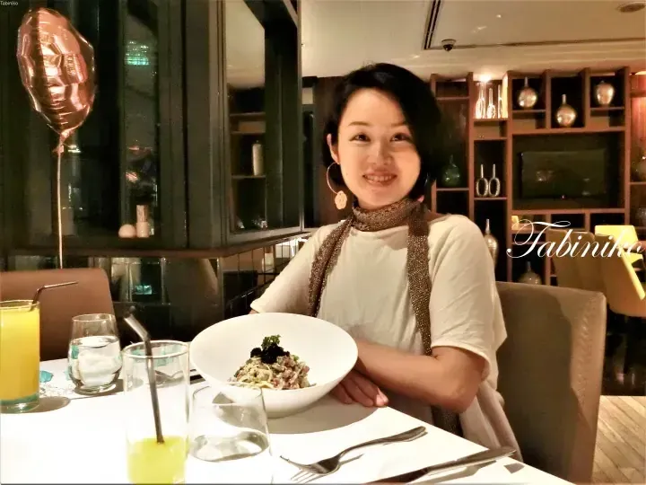 Smiling woman dining at a restaurant table with a bowl of pasta, juice, and a rose gold balloon in the background.