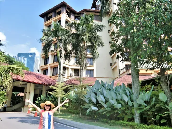 Woman in a straw hat poses with arms outstretched at the entrance of a resort building with palm trees and IOI building in the background.