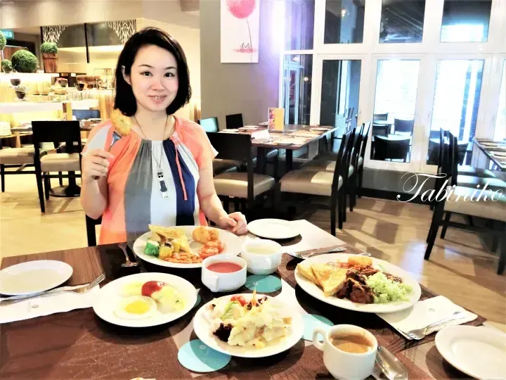 Smiling woman at a breakfast buffet table with plates of eggs, toast, and local dishes in a bright hotel dining area.