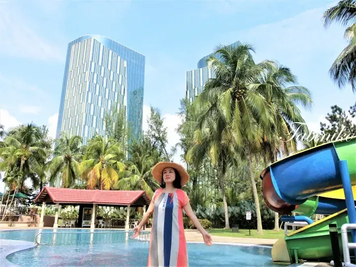 Woman in a colorful dress and straw hat stands by a pool with a water slide, palm trees, and modern high-rise buildings in the background.