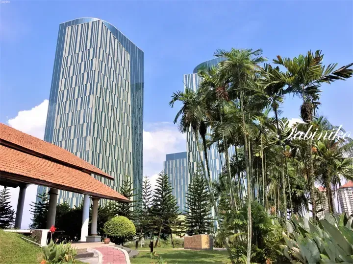 Modern glass skyscrapers rise behind lush tropical palm trees and a traditional pavilion with a terracotta roof.