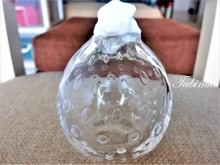Clear glass Christmas bauble with raised dots and white foam on top, resting on a textured beige surface.