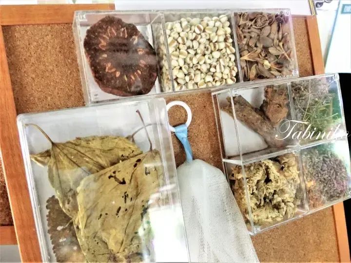 Assorted dried herbs and botanicals displayed in clear plastic containers on a cork board.