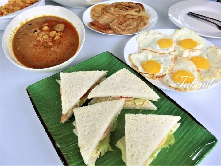 ロティカナイ、目玉焼き、サンドイッチ、カレーが並ぶマレーシアの伝統的な朝食のテーブル。