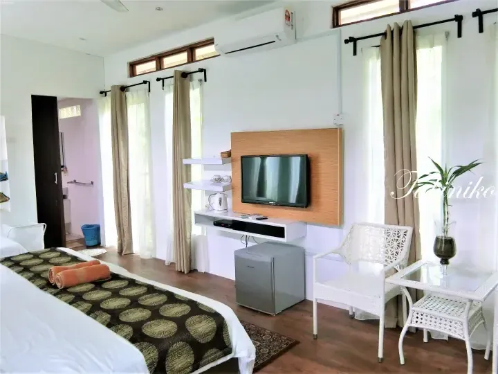 Bright hotel room with a double bed, TV, mini fridge, and white wicker seating area near large windows.