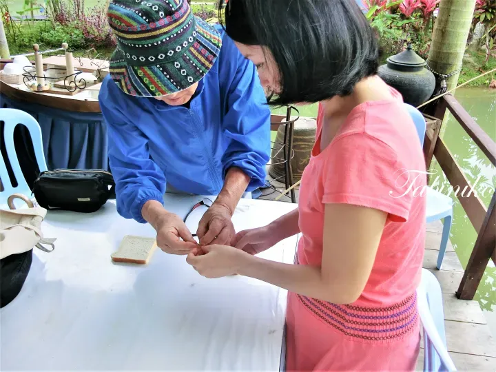 在池塘边的户外桌旁，当地向导正在教游客制作传统手工艺品。