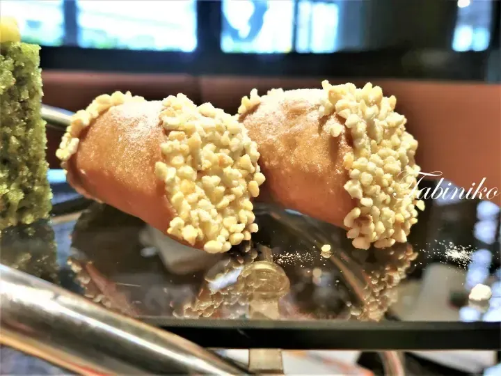 Two golden-brown pastries topped with crumble and powdered sugar displayed on a glass shelf alongside a green cake.