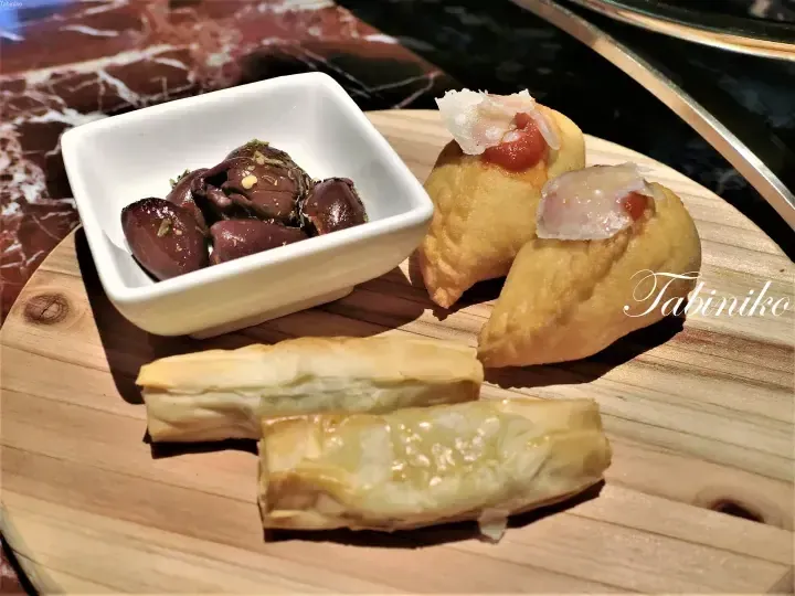 Assorted appetizers including marinated olives, fried dumplings, and pastry rolls served on a wooden board.