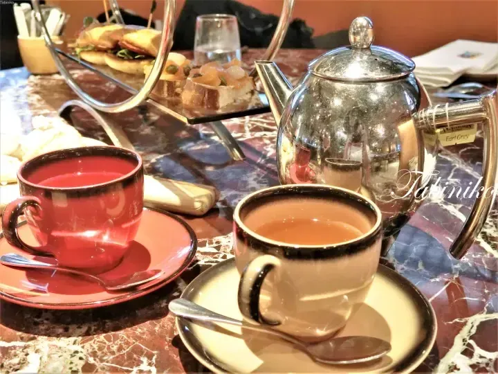 Silver teapot and cups of Earl Grey tea on a marble table with a tiered stand of savory sandwiches and canapés.