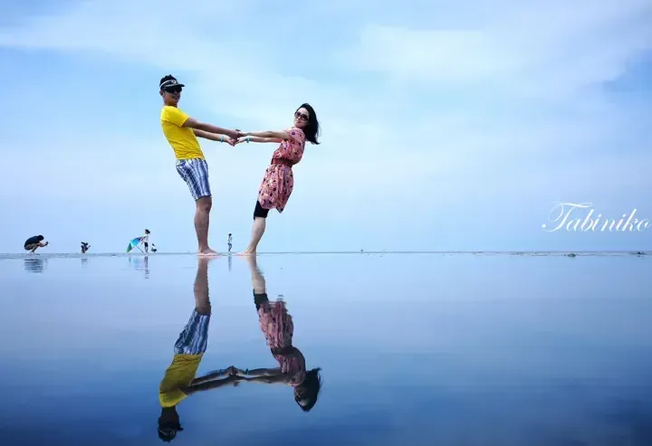 Couple holding hands and leaning back on a reflective salt flat, creating a mirror effect with their reflection.