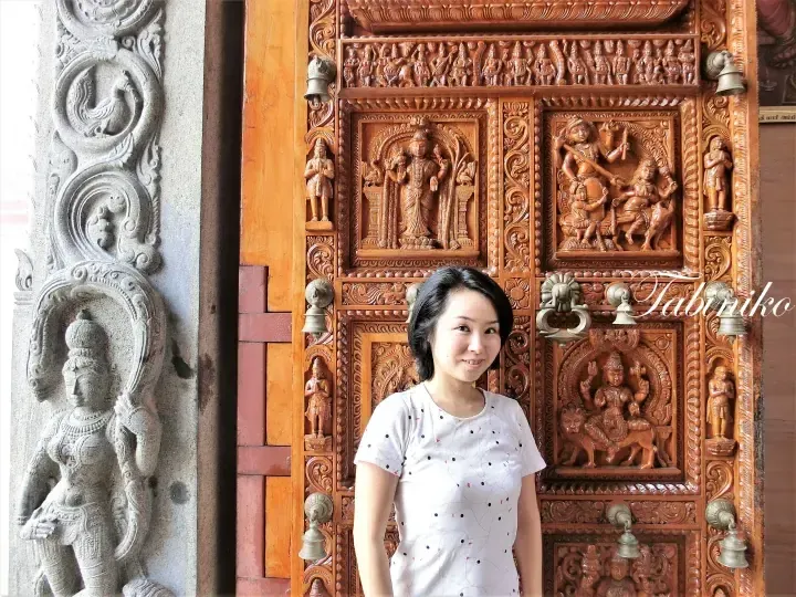 Woman posing in front of an intricately carved wooden temple door with brass bells and a stone deity statue.