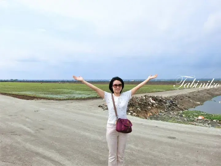 Woman with arms outstretched on a rural road near green rice fields and a canal in Malaysia.