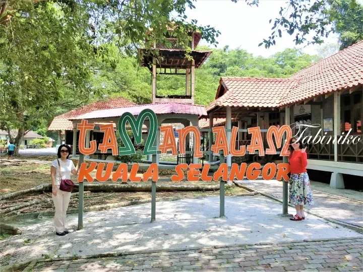 Two women posing beside the Taman Alam Kuala Selangor sign at the entrance of the nature park.
