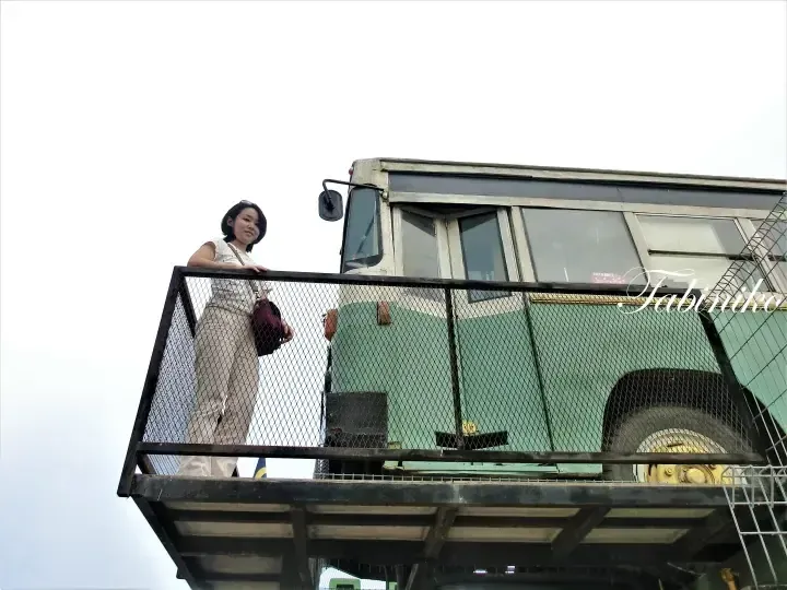 A woman standing on a metal balcony overlooking a large green truck, likely part of a unique property or attraction.