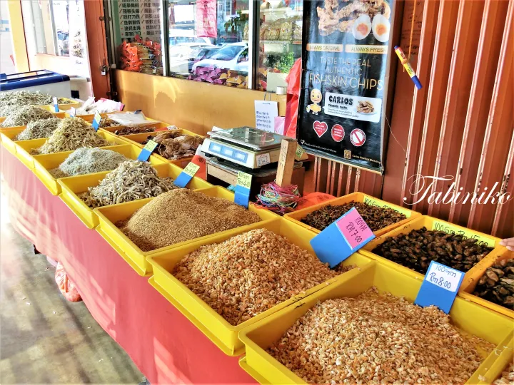 カルロス・フィッシュスキンチップスの看板があるマレーシアの市場で、様々な乾燥魚介類やエビが入った黄色い箱。