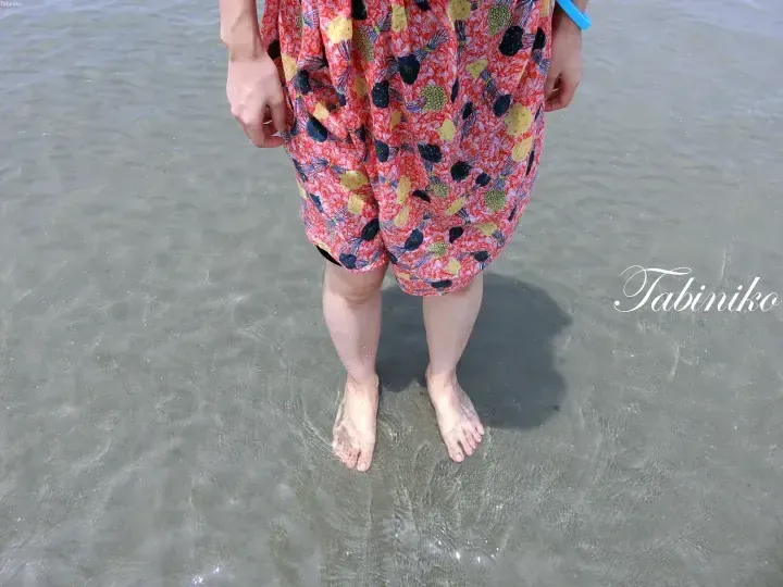 Person standing barefoot in shallow water wearing a red floral dress with a blue wristband.