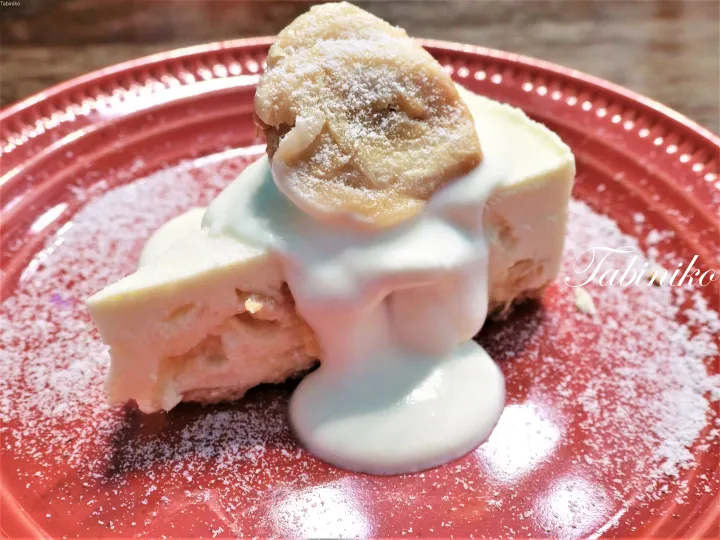 バナナとクリームをトッピングしたバナナチーズケーキの切り身が、粉糖をまぶした赤い皿に盛られている。