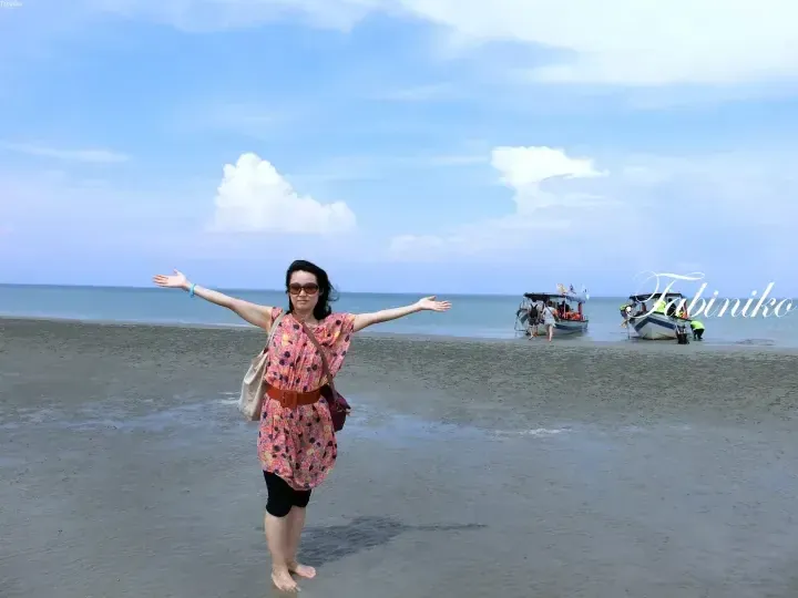 Woman with arms outstretched on a sandy beach with boats in the background.