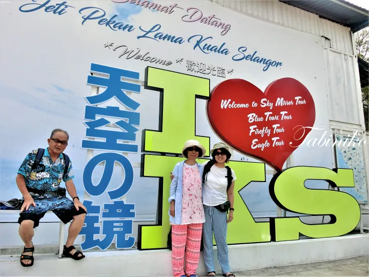 「Selamat Datang Pekan Lama Kuala Selangor」と「Sky Mirror Tour」の看板の前で記念撮影する3人。