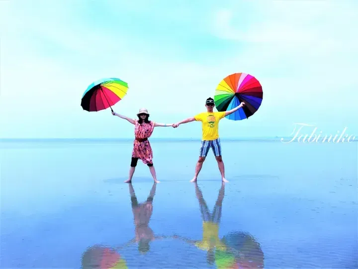 Couple holding rainbow umbrellas and hands on a reflective beach at Tabiniko, Malaysia.