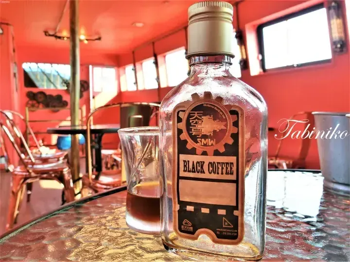 A bottle of Sky Mirror World SMW Black Coffee sits on a table inside a red-themed cafe.