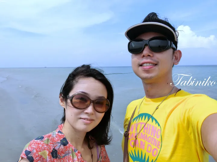 海を背景に砂浜に立つ、サングラスと帽子をかけたカップルの笑顔のポートレート。
