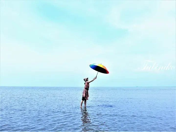 Woman holding a rainbow umbrella while standing in the ocean water under a clear blue sky.