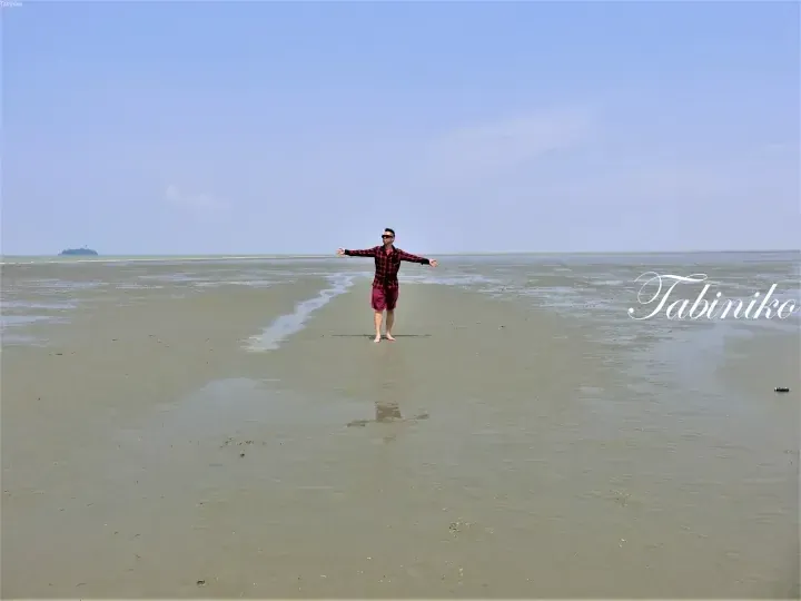 Man standing on vast mudflats with arms outstretched near a distant island under a clear blue sky.