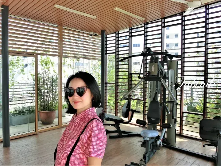 Woman in sunglasses and red checkered shirt poses in a bright gym with wooden slat walls and fitness equipment.