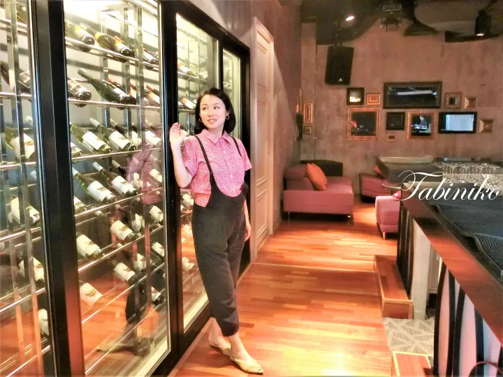 ピンクのシャツとオーバーオールを着た女性が、木製床とピンクのソファがあるラウンジでワインセラーの隣に立っている。