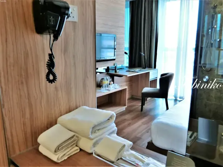 Modern hotel room interior with neatly folded towels, a wall-mounted hairdryer, and a desk area near a window.