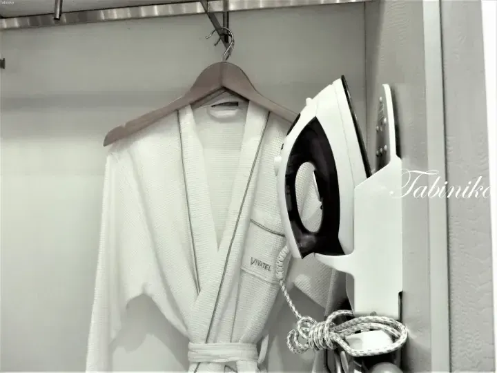 White waffle bathrobe with VIVATEL logo hanging on a wooden hanger next to an iron in a hotel closet.