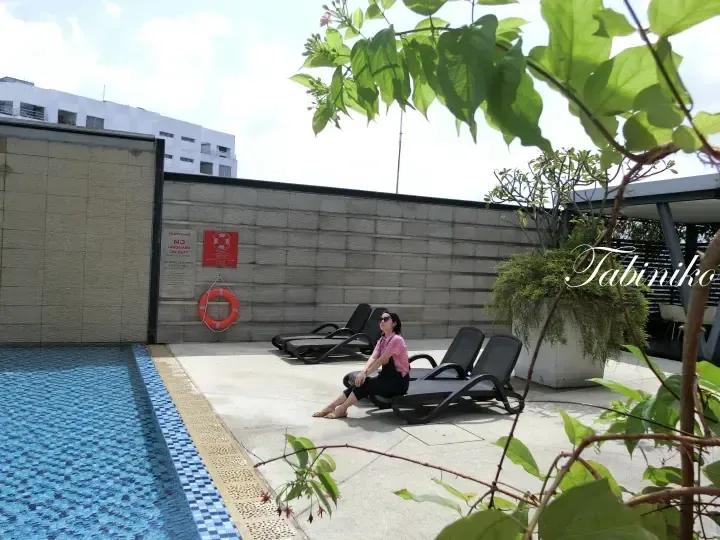 Woman relaxing on a sun lounger beside a rooftop pool at a Malaysia property, with safety signage and greenery visible.