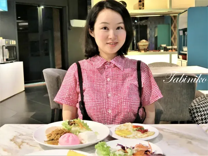 Smiling woman in a pink checkered shirt sits at a dining table with plates of breakfast food including noodles, buns, and omelets.