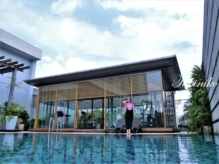 Woman posing by a rooftop pool beside a modern glass-walled gym with wooden louvers.