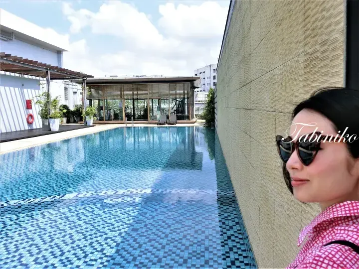 A woman in sunglasses stands beside a rooftop swimming pool with blue tiles, overlooking a modern building and gym area.