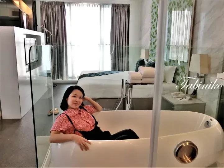 Woman relaxing in a freestanding bathtub within a modern hotel suite featuring a bed and large window.