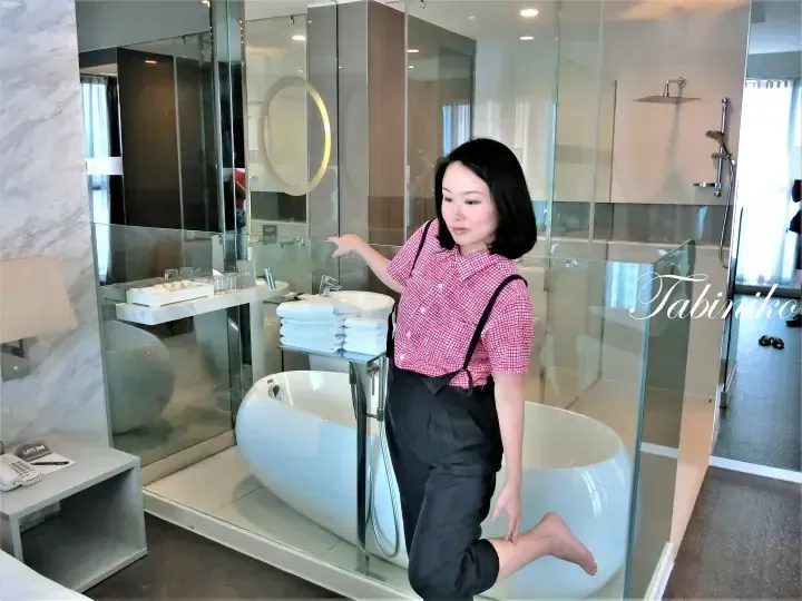 Woman in pink checkered shirt poses beside a modern white freestanding bathtub in a luxury hotel bathroom with glass partitions.