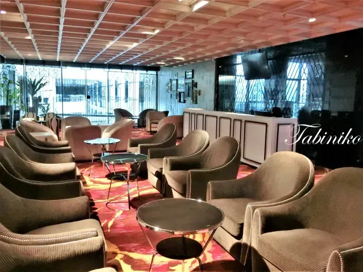 Elegant lounge area with plush brown armchairs, red patterned carpet, and decorative ironwork windows at a luxury hotel.
