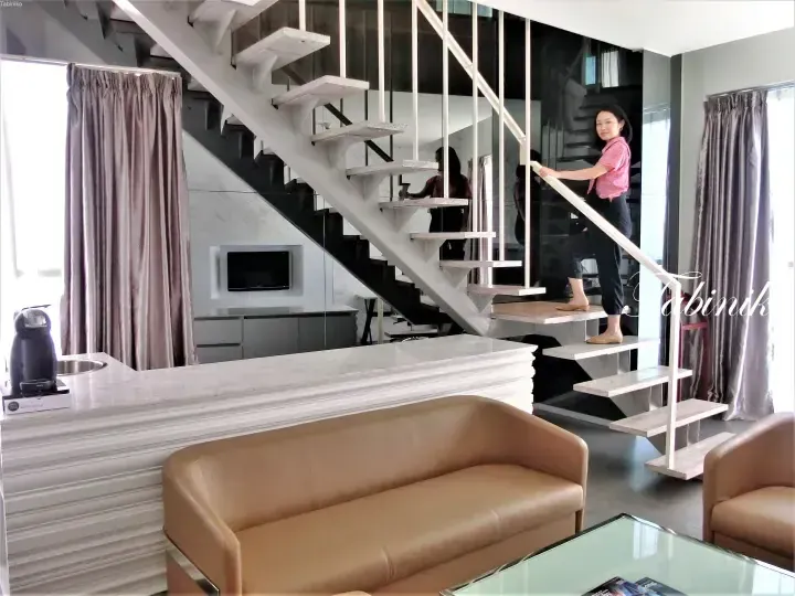 Woman ascending a modern floating staircase inside a stylish loft apartment featuring a beige leather sofa and marble kitchen counter.