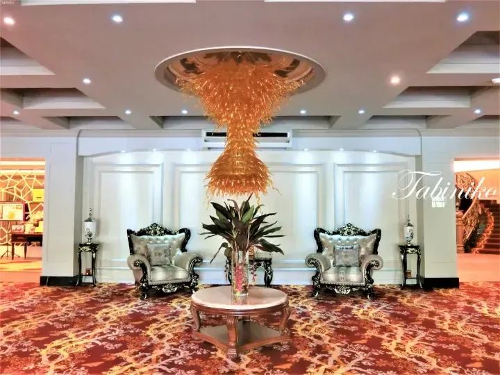 Elegant hotel lobby with ornate chairs, a large orange chandelier, and patterned carpet.