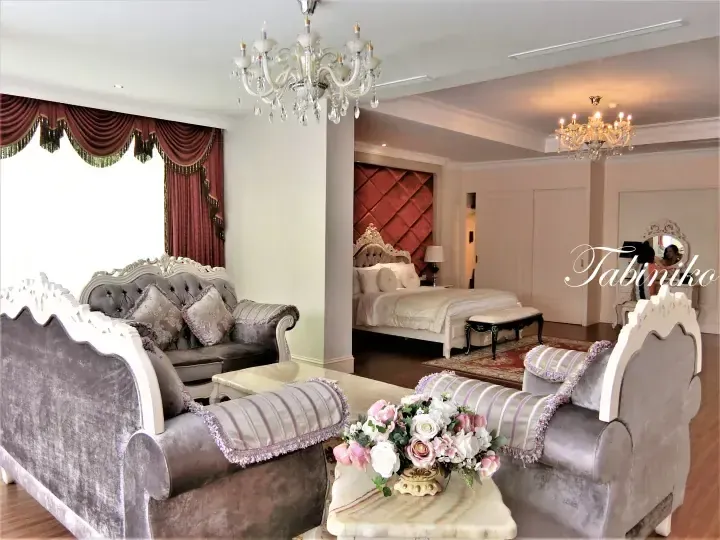 Luxurious hotel suite interior featuring a living area with grey velvet sofas and a bedroom with a tufted red headboard.