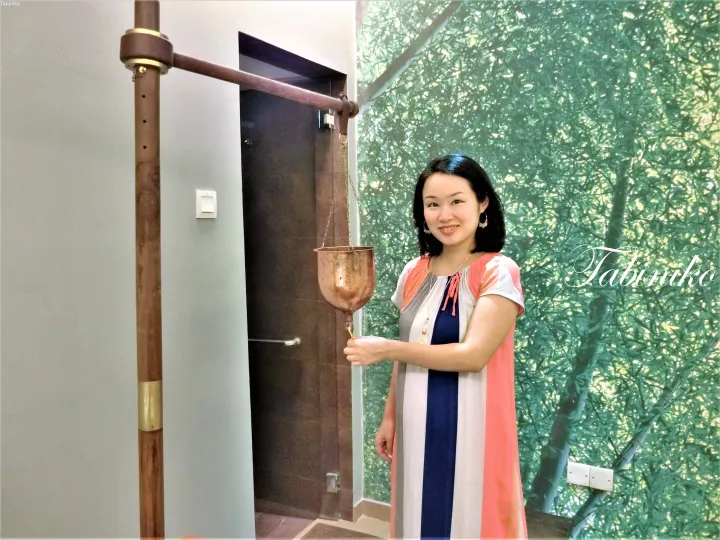自然をイメージしたバスルームで、木製のシャワー設備で伝統的な銅の柄杓を持つ女性。