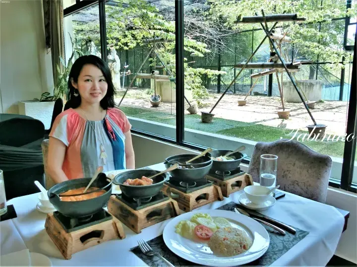 Woman dining at a table with multiple hot pots and rice, overlooking a lush bird aviary enclosure.