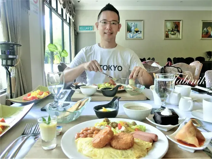 Man eating breakfast at a table filled with omelets, pastries, and fruit at a Malaysian hotel restaurant.