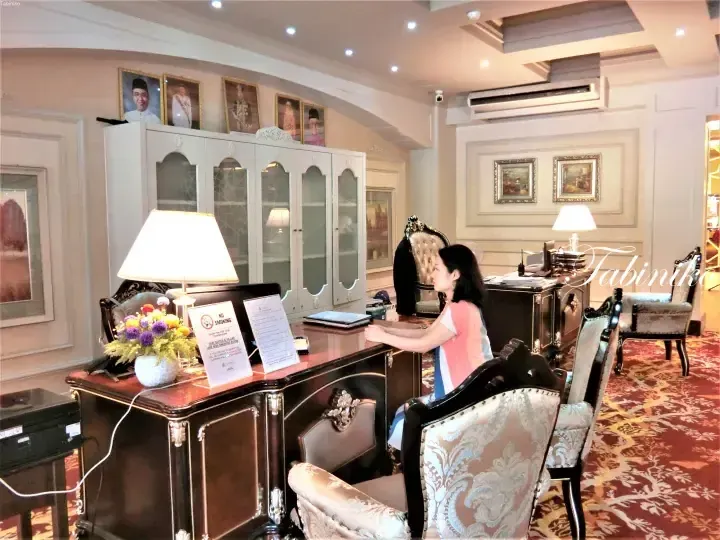 A woman sits at a dark wood reception desk in a hotel lobby, with a &ldquo;No Smoking&rdquo; sign, floral arrangement, and framed portraits on a white cabinet.