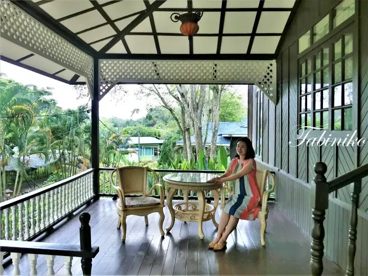 Woman relaxing on a wooden veranda overlooking lush tropical greenery at a Malaysian property.